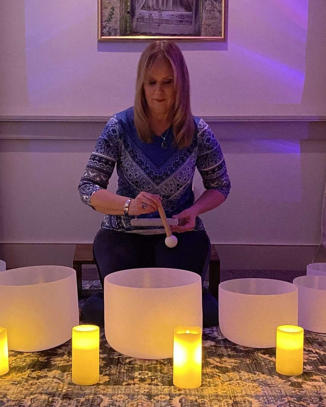 A picture of Clarissa Wrightson sitting in front of a semi circle of Crystal Singing Bowls ready to play. Her space is warmly lit with a beautiful persian rub on the floor and purple and green mood lighting reflected on the walls behind her.