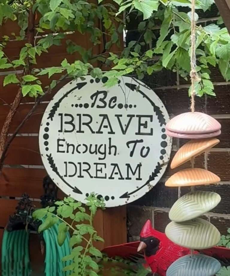 Garden sign reading “Be Brave Enough to Dream” surrounded by greenery and colorful garden decorations.