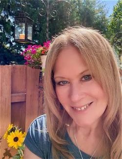 Portrait of Clarissa Wrightson, Reiki Master and Energy Helaer in a garden surronded by flowers with a fence and trees beyond in the background. There is also a bird house over her left shoulder